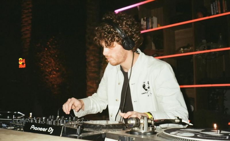 DJ with curly hair wearing headphones and a white jacket, focused on mixing tracks on a Pioneer DJ deck in a dimly lit venue.