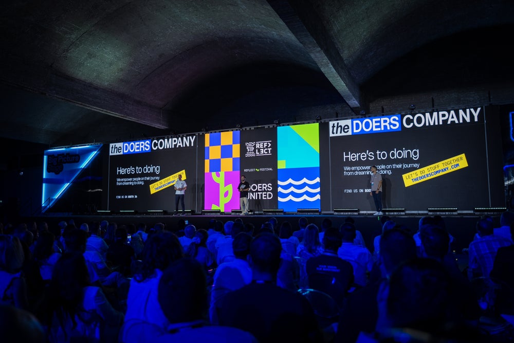 A packed conference hall at Doers Summit where speakers stand on a wide stage in front of large colourful screens branded “the DOERS Company” as the audience watches.