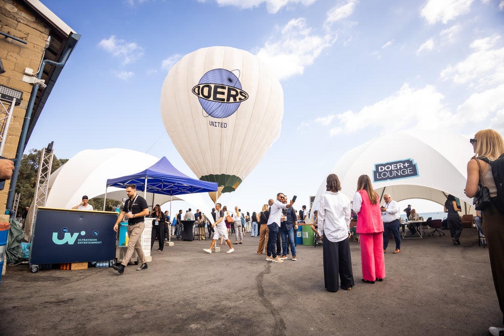 An outdoor networking area at Doers Summit with attendees mingling around tents and a large white balloon marked “DOERS United” against a blue sky.