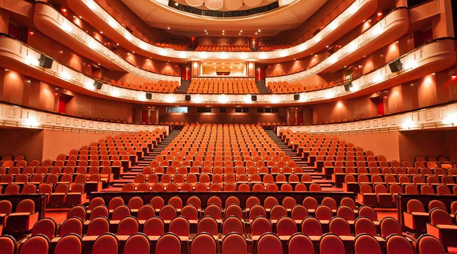 Main hall of the Cairo Opera House with rows of red upholstered seats and curved balconies glowing in warm light, ready to welcome audiences for upcoming performances. القاعة الرئيسية في دار الأوبرا المصرية، تضم صفوفًا من المقاعد الحمراء المنجدة وشرفات منحنية مضيئة بضوء دافئ، استعدادًا لاستقبال الجماهير في العروض القادمة.
