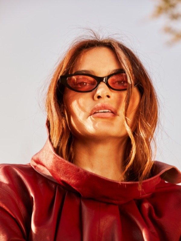 Woman in a red leather hooded top and tinted cat-eye sunglasses, lit by warm sunlight with loose waves framing her face