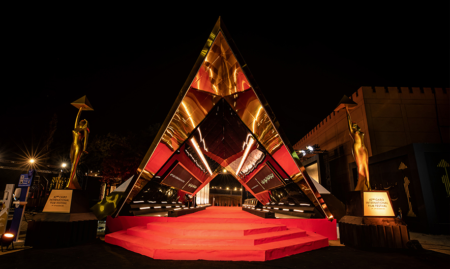 Illuminated pyramid-shaped entrance and red-carpet stairs at the Cairo International Film Festival at night, flanked by two golden statuettes