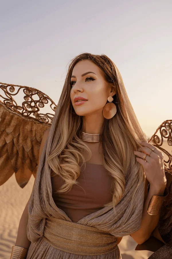 Woman in a gold-toned gown with sculpted wings posing in the desert at sunset, styled with statement earrings and layered metallic jewellery.