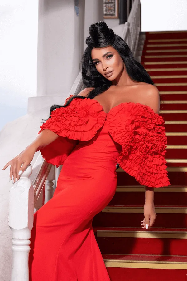 Woman in a dramatic off-the-shoulder red gown with large ruffled sleeves, leaning on a staircase railing in front of red-carpeted steps.