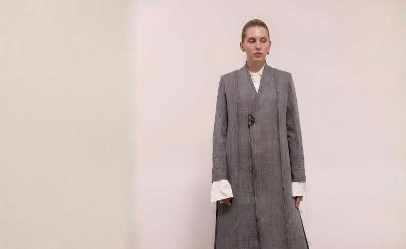 Woman in a long, tailored grey coat layered over a white shirt, standing against a minimal light background in a neutral monochrome look