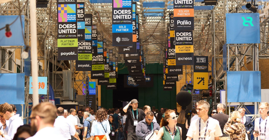A busy outdoor walkway at Doers Summit with crowds of attendees networking beneath a canopy of sponsor banners and signs reading “Doers United.”