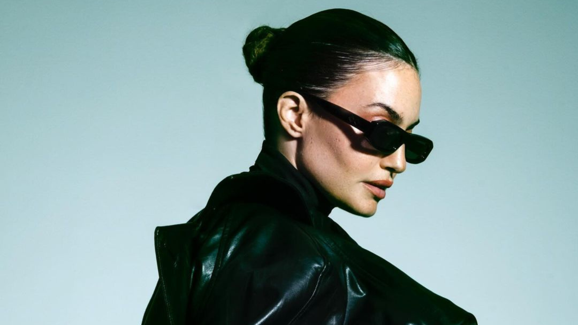 Woman in a sleek black leather jacket wearing narrow rectangular sunglasses, hair pulled back in a bun, posing in profile against a soft green background.”