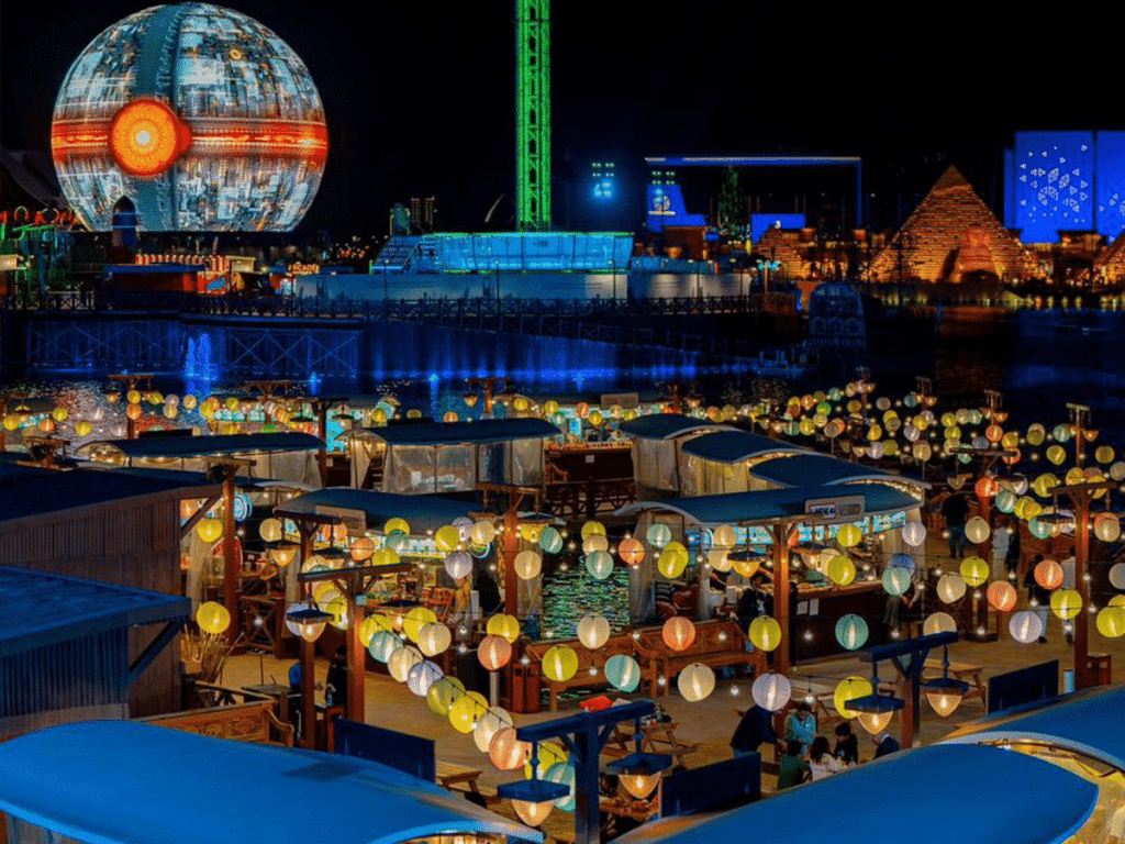 Lantern-lit waterfront marketplace at Riyadh Season with colourful hanging lights, stalls and visitors, set against a backdrop of futuristic rides including a huge glowing spherical structure.