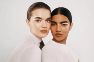 Two models wearing minimalist white tops and graphic white eyeliner pose together against a clean studio background, representing modern clean beauty and diversity عارضتان ترتديان قمصانًا بيضاء بسيطة وماكياج عيون أبيض بخطوط جرافيكية، تتخذان وضعية أمام خلفية استوديو نظيفة، في تمثيل للجمال النقي العصري والتنوع.