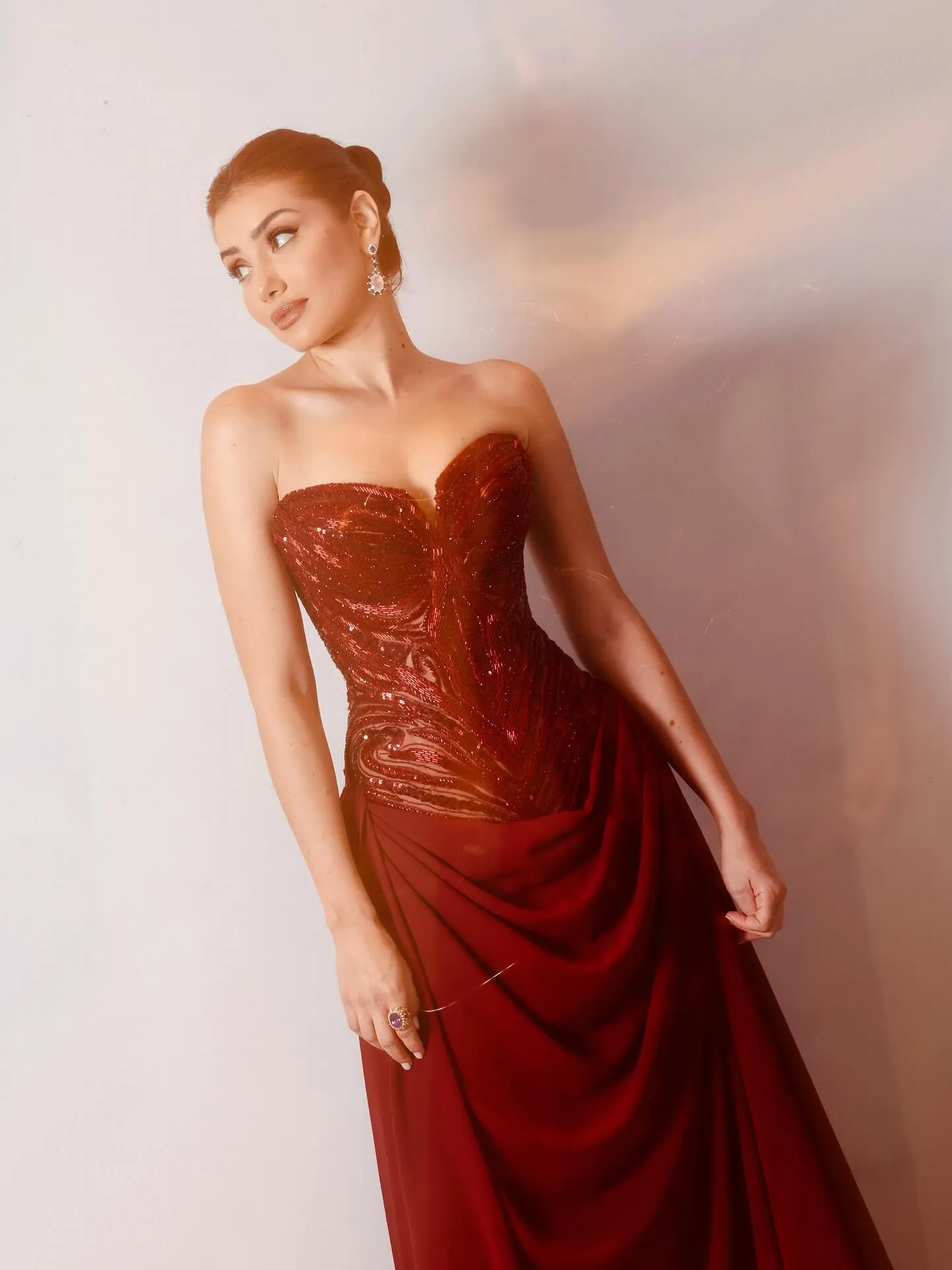 Elegant Egyptian actress in a strapless, ruby-red embellished gown, posing against a soft neutral backdrop with her hair in a sleek bun and sparkling drop earrings completing the red-carpet look.