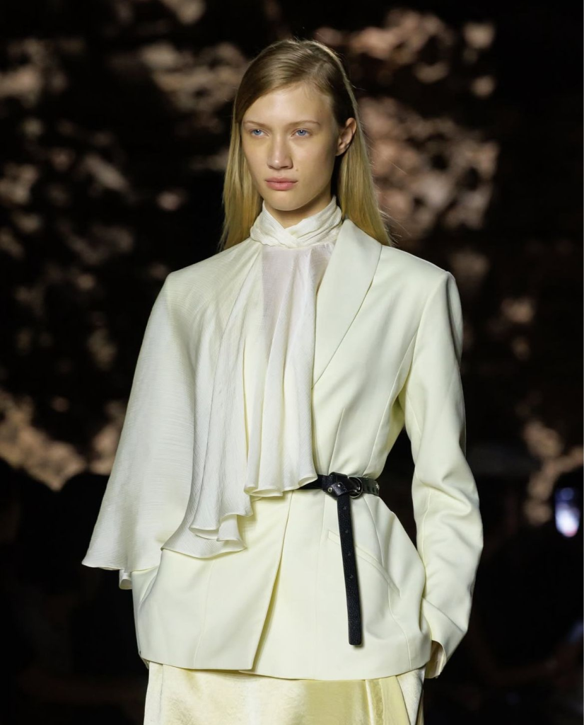 Riyadh Fashion Week 2025: From Local Talent to Vivienne Westwood’s Middle East Debut 2 2 Model on the catwalk in a soft cream tailored blazer layered over a sheer high-neck blouse, cinched at the waist with a slim black belt.