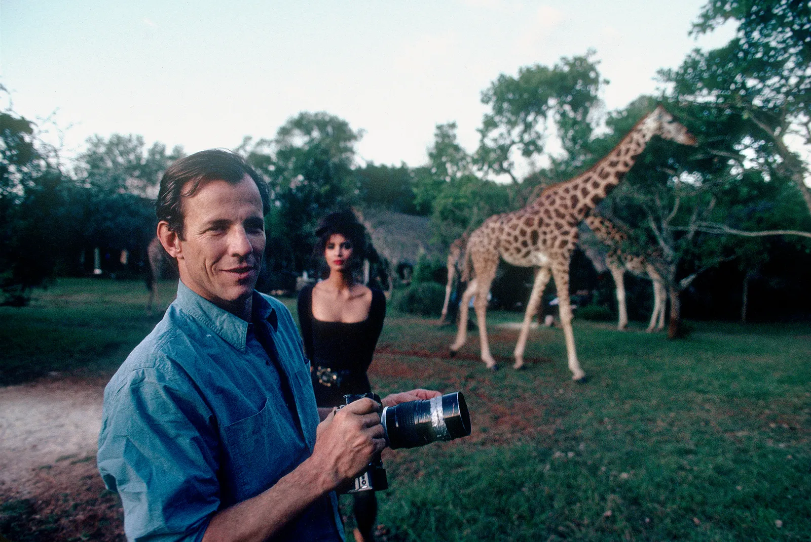 Man in a blue shirt holding a camera in an open grassy landscape, with a woman standing behind him and giraffes walking among the trees in the background رجل يرتدي قميصًا أزرق يحمل كاميرا في منظر طبيعي عشبي مفتوح، وخلفه تقف امرأة بينما تسير الزرافات بين الأشجار في الخلفية