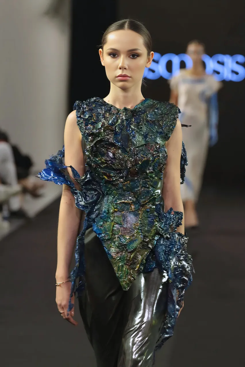 Young woman in a green, embellished gown standing against a plain studio backdrop, her hair in sleek bun
