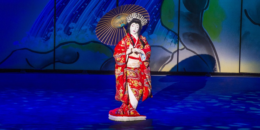 Japan Festival Riyadh 2025: Anime, Food and Diplomacy 1 1 A performer in an ornate red kimono and white makeup holds a paper parasol on a blue-lit stage, evoking traditional Japanese theatre during a cultural show.