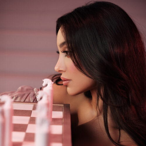 Zaza Bajouk Is the Middle East’s #1 Fashion Icon in 2025 2 2 Side-profile portrait of a woman with long dark hair, lit in pink tones, leaning close to a pink-and-white chessboard with stylised chess pieces in the foreground.