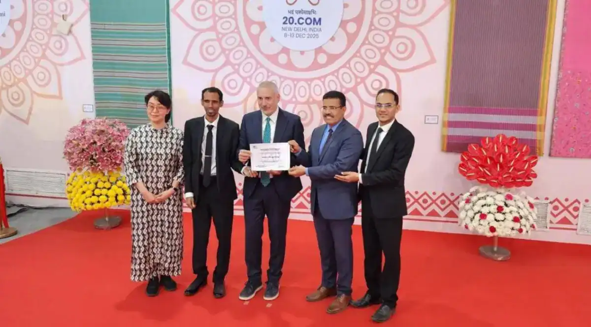 Group of officials on stage holding a certificate during a cultural recognition ceremony, with decorative textiles and floral arrangements in the background.