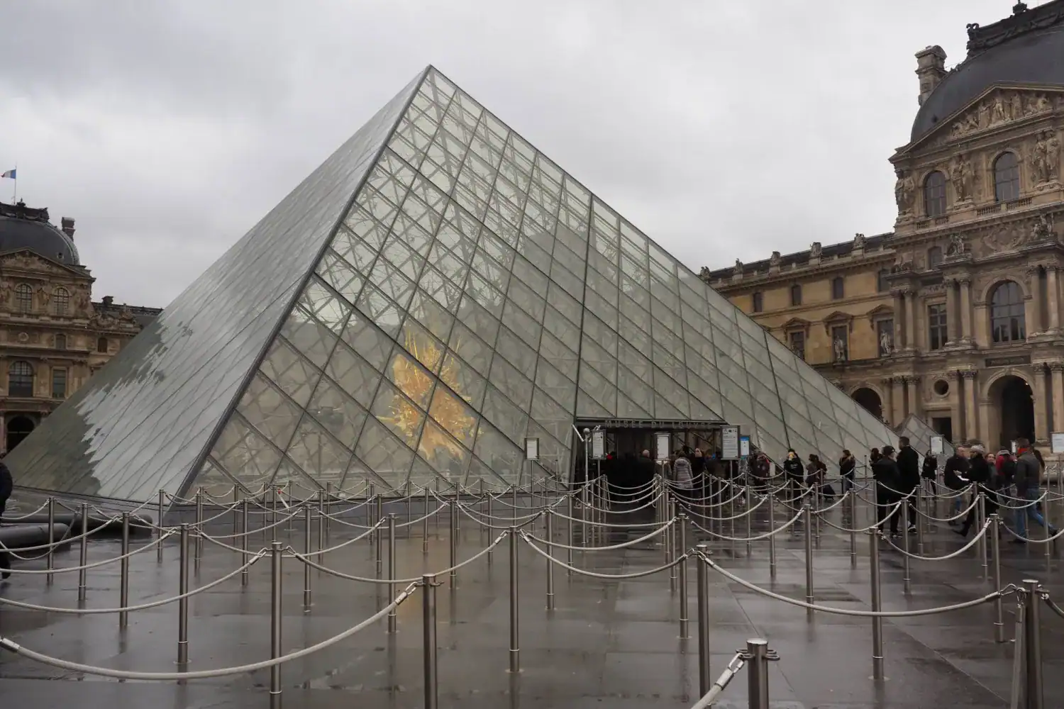 The glass pyramid at the Louvre Museum in Paris with empty queue barriers and visitors gathered outside the closed entrance.