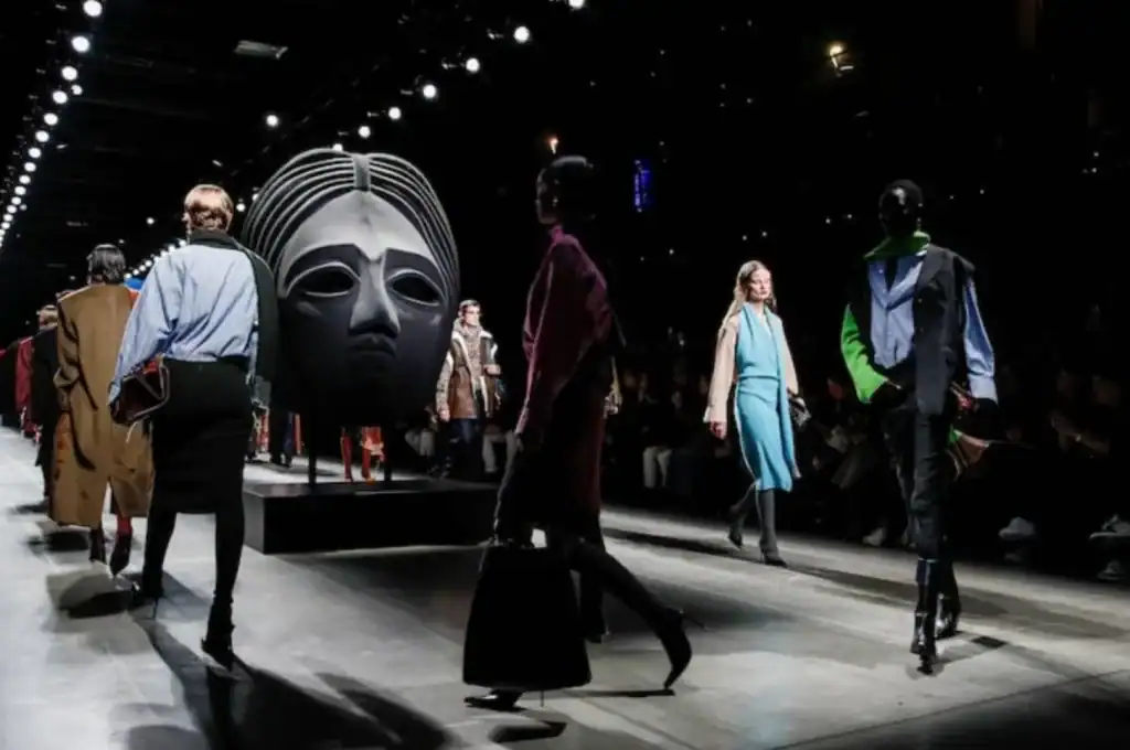 Models walk a runway during a fashion show featuring a large sculptural art installation at the center of the catwalk.