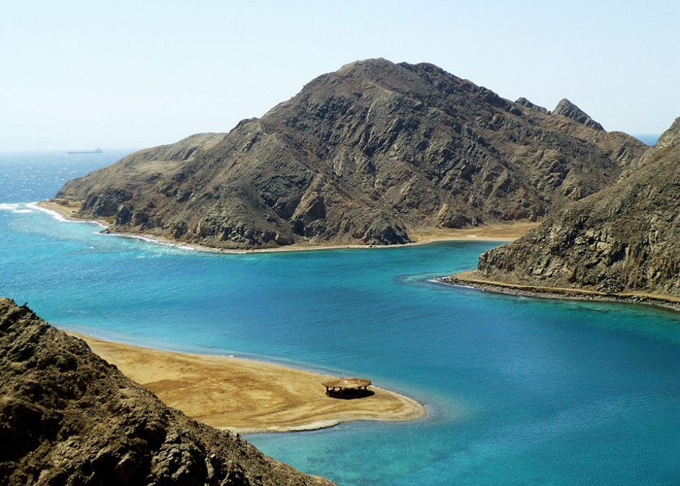 Coastal landscape in North Sinai showing a turquoise lagoon bordered by rugged mountains and a sandy shoreline under a clear sky.