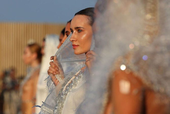 Model wearing a sheer, textured garment during an outdoor fashion presentation, with other models softly blurred in the background.