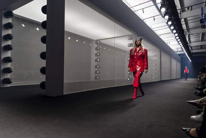 Model walking down a runway wearing a red tailored outfit during a fashion show, with seated guests along the catwalk.