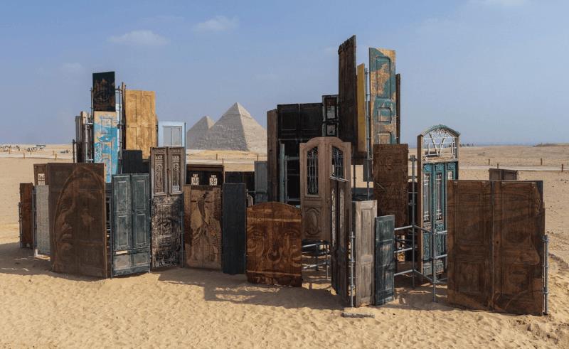 Outdoor art installation composed of reclaimed wooden doors arranged in the desert landscape near the Giza pyramids, forming Vhils’ “Doors of Cairo” installation.