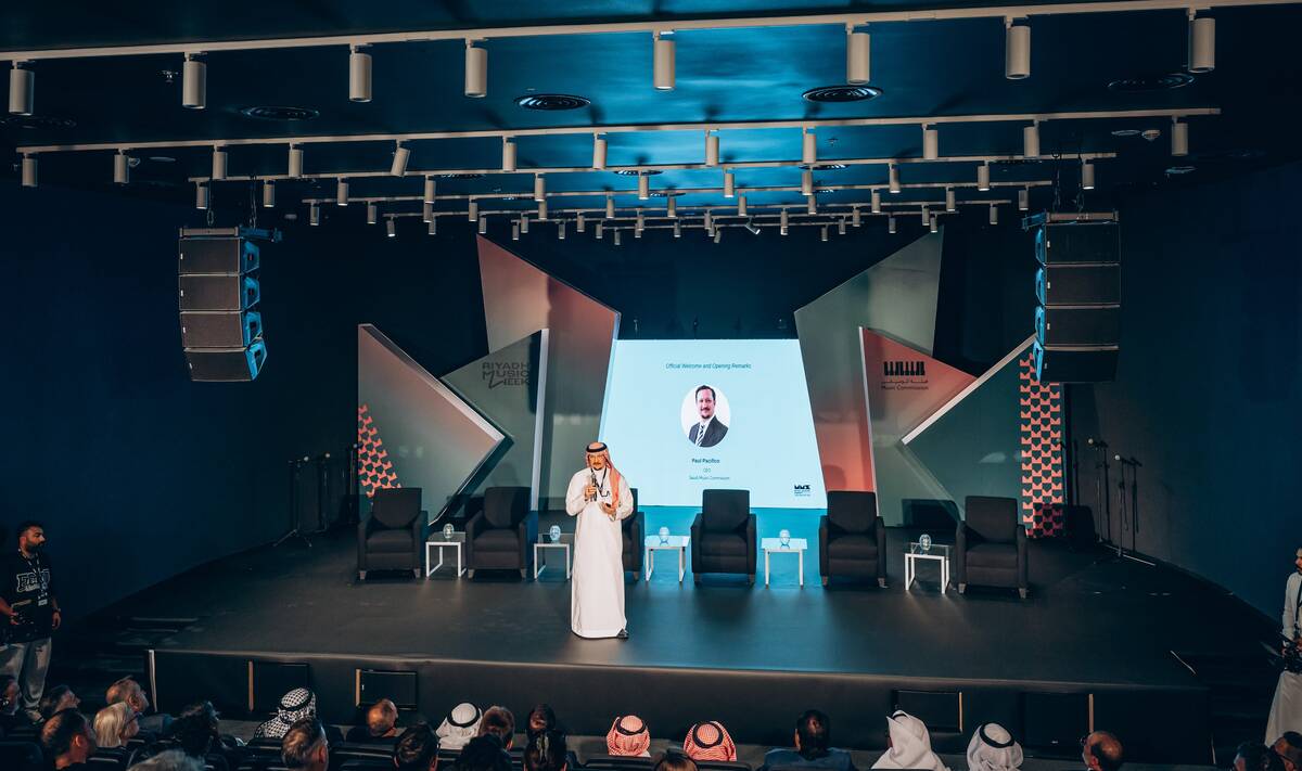 Speaker in traditional Saudi attire presenting on a modern conference stage with a large screen backdrop, seated panel chairs, and an audience attending a business or innovation event.