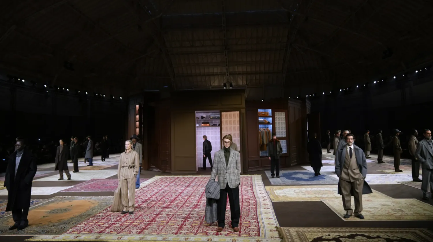 Wide view of a menswear runway set inside a dark hangar-like venue, with models standing spaced apart on large patterned rugs around a central room-like installation.