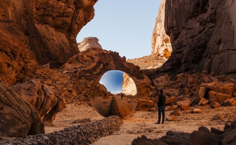 A mirrored spherical sculpture sits in a rocky desert canyon, reflecting the surrounding sandstone cliffs as a visitor stands nearby.