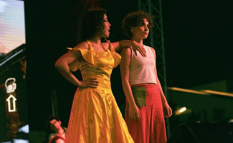 Egypt Launches National Initiative to Revive Arts & Culture in Universities 3 3 Two performers stand under stage lighting during a live performance, one in a yellow dress and the other in a white top with a patterned skirt.