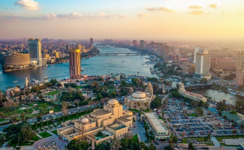 Aerial view of central Cairo at sunset, with the Nile River winding through the city skyline and the Cairo Opera House complex visible in the foreground.