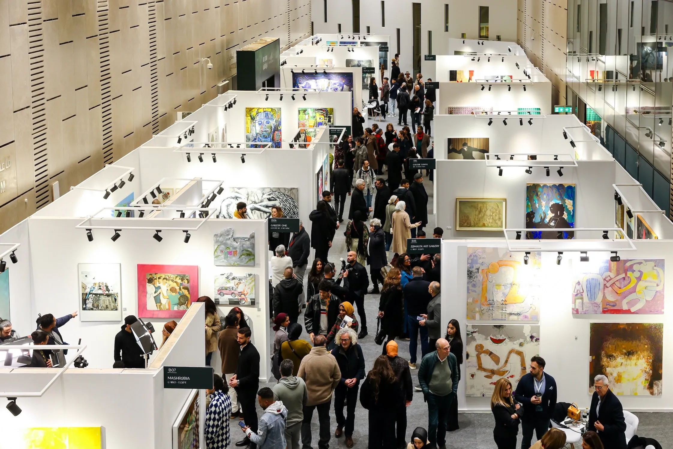 Overhead view of Art Cairo exhibition halls with white gallery booths, colorful artworks on the walls, and crowds moving through the central aisle.