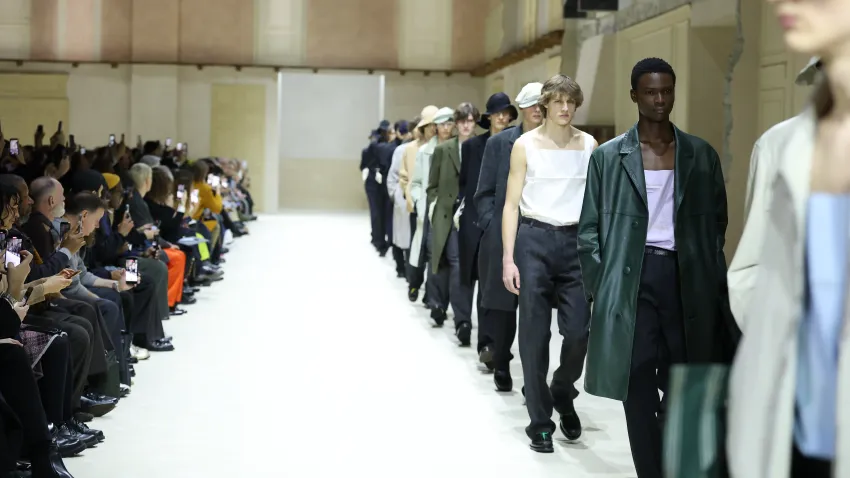 Menswear runway finale with a line of male models walking toward the camera on a bright white catwalk, while seated guests along the side film and watch.