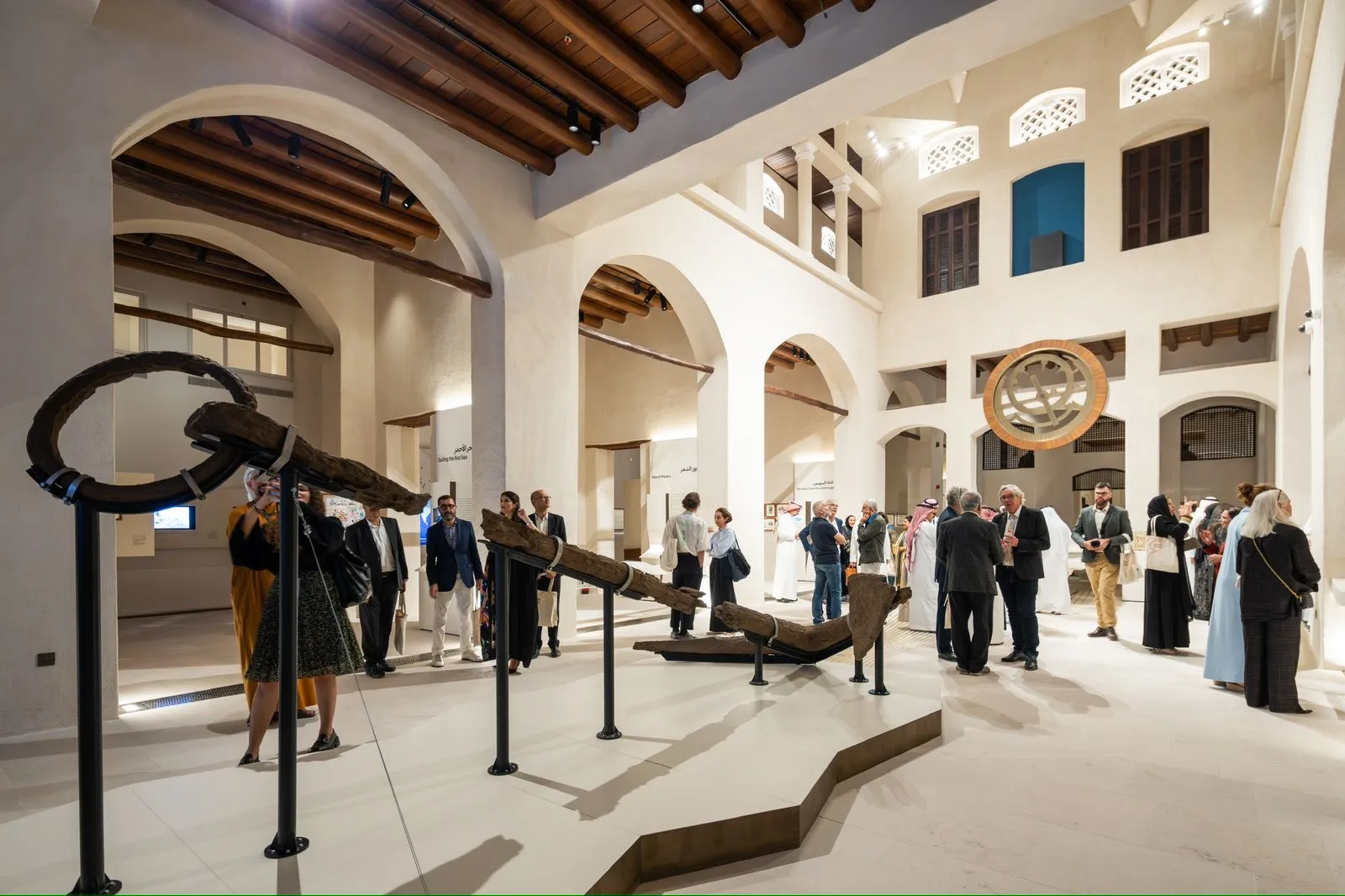 Interior view of a museum courtyard with arched walkways, wooden ceiling beams, visitors gathered around exhibits, and a large suspended anchor installation in the center.