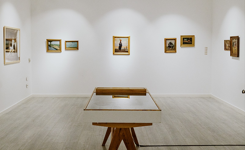 Wide view of a minimalist gallery room with multiple small framed paintings on white walls and a glass display case centered in the space.
