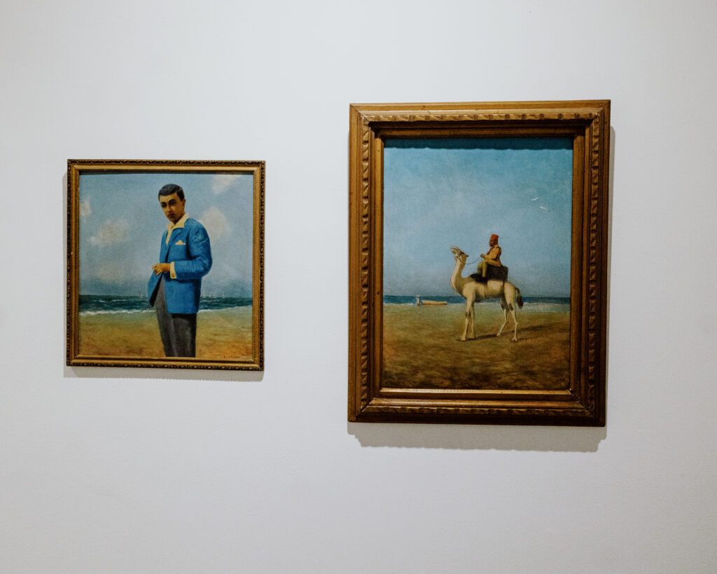 Two framed paintings on a white gallery wall: a portrait of a man in a blue jacket by the sea and a scene of a rider on a camel against a wide sky.