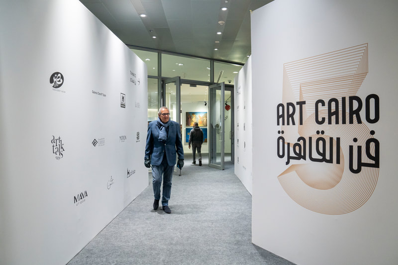 Art Cairo entrance corridor with white walls, partner logos on the left, and a large “ART CAIRO / فن القاهرة” graphic on the right, with visitors walking inside.