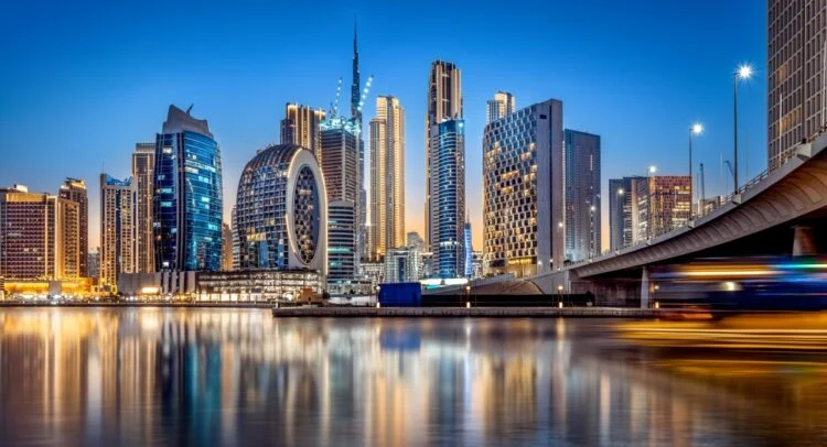 Night skyline of Dubai’s Business Bay with illuminated high-rise towers reflecting on calm water beside a modern bridge, with light trails from passing traffic.