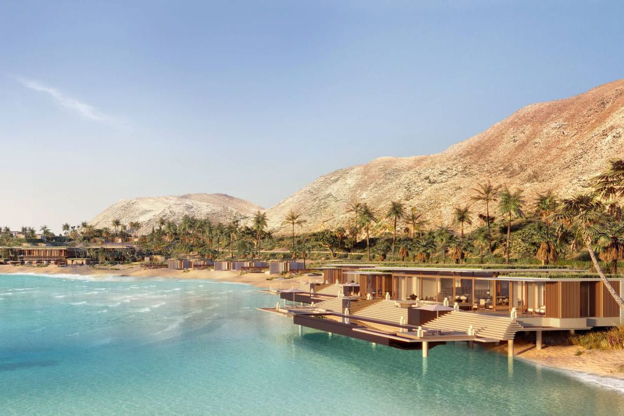 Luxury beachfront resort with modern wooden villas extending over turquoise water, set against palm-lined shores and sunlit desert mountains in a serene coastal landscape