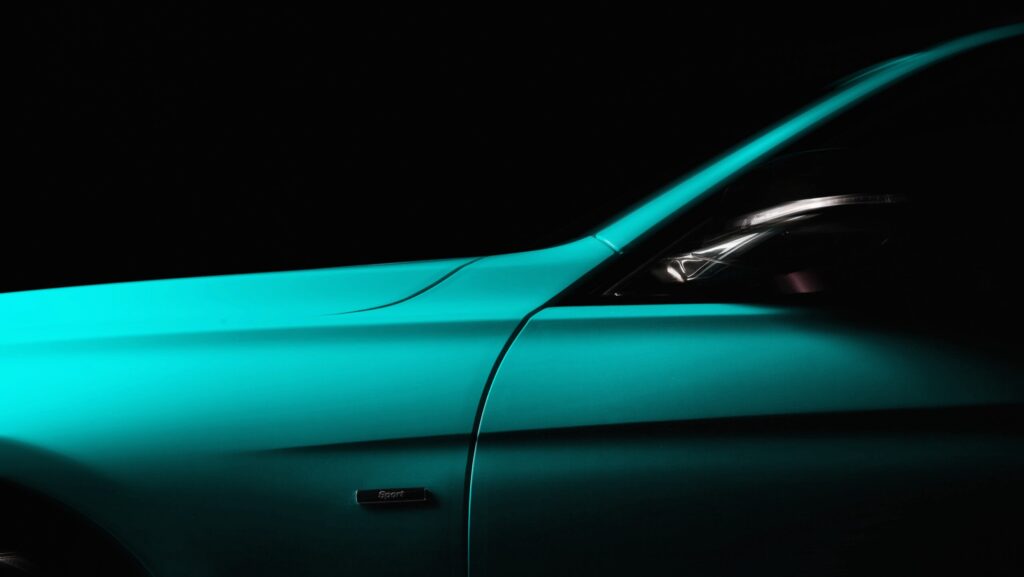 Close-up of a sleek teal luxury car side profile in low light, highlighting smooth body lines, side mirror, and modern headlight design against a dark background.