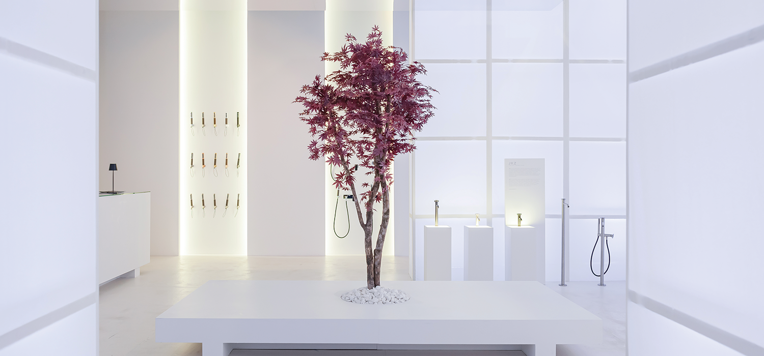 Minimal white bathroom exhibition space with a sculptural tree centerpiece, soft lighting, and contemporary fixtures, reflecting Salone del Mobile’s focus on wellbeing, design innovation, and sensory living.